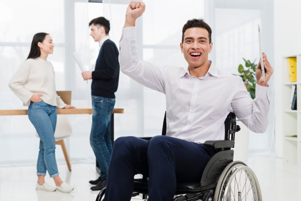 A participant in a wheelchair cheerfully raises his fist, holding a tablet, in a bright office. Two support worker chat in the background, conveying teamwork.